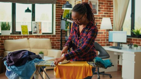 Photo for African american housewife using iron on ironing board to do housekeeping work, casual adult ironing clothes in living room. Young woman smoothing out garment and clothing, chores. - Royalty Free Image