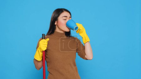 Foto de Cheerful housekepper enjoying cup of coffee while cleaning house with broom, standing in studio over blue background. Maid using protective equipment and sanitary practices to keep customers safe. - Imagen libre de derechos