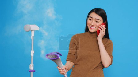 Foto de Cleaning lady talking at mobile phone with remote friend while using steamer to ironing clothes in studio over blue background. Woman used the best cleaning products and equipment - Imagen libre de derechos