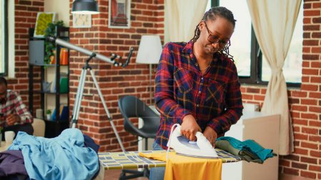 Foto de Depressed african american woman ironing laundry, getting angry at husband not helping with house chores. Tired stressed girlfriend taking care of household by herself, washed garment. - Imagen libre de derechos