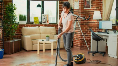 Téléchargez les photos : Young woman sweeping dirt with mop in living room, mopping wooden floors and cleaning apartment. Casual female person washing tiles with cleaner, being focused on housework or chores. - en image libre de droit