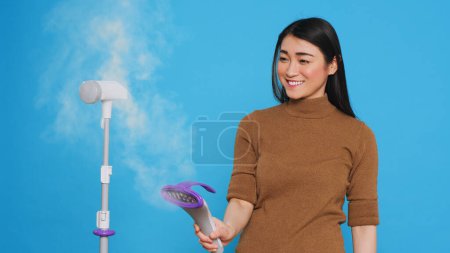 Photo for Smiling cleaning lady using steamer to ironing clothes in studio over blue background. Woman used the best cleaning products and equipment to ensure a sparkling, fresh-smelling home for her clients. - Royalty Free Image