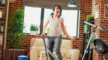 Foto de Happy housewife listening to music on headphones and vacuuming floors, having fun with vacuum cleaner. Young smiling person feeling positive while she does spring cleaning at home. Handheld shot. - Imagen libre de derechos