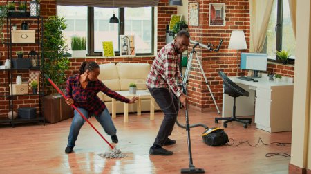 Foto de African american couple dancing and cleaning apartment rooms, using mop to wash dirt and vacuum to clean floors. Young life partners enjoying spring cleaning with washing solution. - Imagen libre de derechos