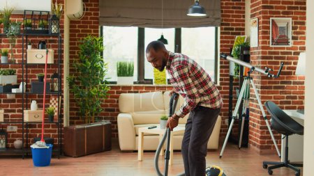 Foto de African american partner using vacuum to clean dust in living room, doing spring cleaning work together at home. Young man vacuuming apartment floors and helping with housework. - Imagen libre de derechos