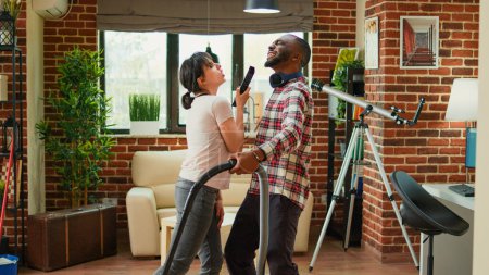 Photo for Diverse husband and wife dancing and sweeping floors, using vacuum cleaner and all purpose cleaner. Happy man and woman laughing and enjoying spring cleaning at home, household chores. - Royalty Free Image