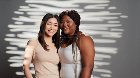 Téléchargez les photos : Happy interracial beautiful women posing together in studio, showing self confidence and body acceptance. Two confident skincare models hugging and promoting body and skin diversity. - en image libre de droit
