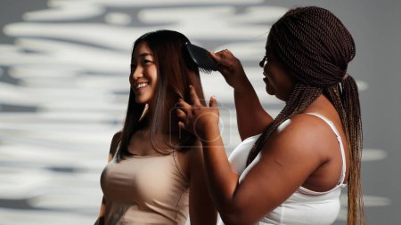 Foto de Persona segura cepillando el cabello de la mujer asiática, usando cepillo para peinar y expresando el concepto de aceptación corporal. Modelos positivos de belleza posando para campaña publicitaria de cuidado de la piel, diversidad de tipos de cuerpo. - Imagen libre de derechos