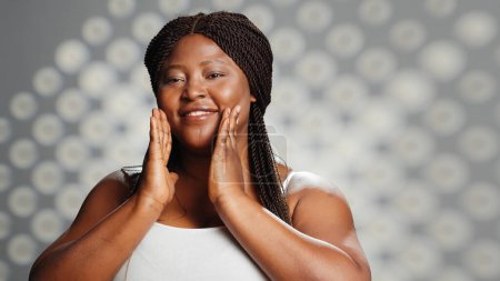 Foto de Chica afroamericana que promueve la crema hidratante en el estudio, la aplicación de suero y crema facial para anunciar la campaña de cuidado de la piel. Modelo natural impecable usando cosméticos dermatológicos, productos. - Imagen libre de derechos