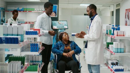 Foto de Social worker helping wheelchair user to buy medicine from pharmacy, receiving advice from specialist. Young adult in wheelchair talking to pharmacist about prescription treatment. - Imagen libre de derechos
