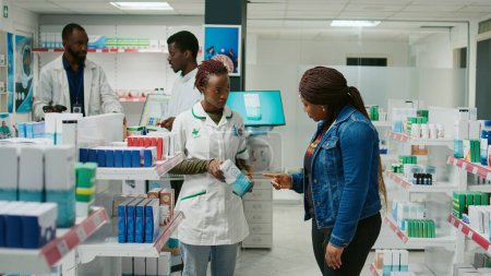 Téléchargez les photos : Female pharmacist helping customer with medicine, showing pharmaceutical products from drugstore shelves. Young adult asking health specialist about boxes of pills and supplements. - en image libre de droit