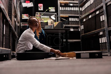 Photo for Private detective studying investigation reports on floor in agency office room. Woman police investigator reading crime case file and analyzing clues sitting with crossed legs - Royalty Free Image