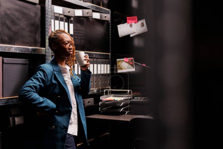 Photo for Concerned african american woman detective drinking coffee and thinking about crime case. Thoughtful criminologist holding tea mug and analyzing evidence in investigator room at night - Royalty Free Image