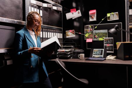 Foto de Una mujer afroamericana investigadora de la policía leyendo el archivo del caso en la oficina. Detective privado que sostiene la carpeta y analiza el informe de investigación cerca del tablero con el esquema de conexión de pistas - Imagen libre de derechos