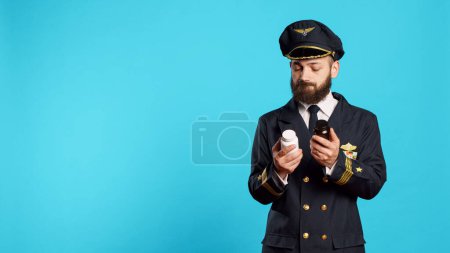 Photo for Male aviator looking at bottles of pills in studio, feeling unsure about medicaments and medical treatment. Young adult flying pilot holding jars of medicine and supplements, aviation worker. - Royalty Free Image