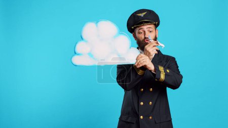 Téléchargez les photos : Aviateur masculin jouant avec un jouet d'avion et un tableau nuage, tenant une icône en carton et un petit avion miniature sur la caméra. Jeune pilote aux commandes avec uniforme aviation publicité transport aérien. - en image libre de droit