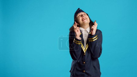Photo for Positive air hostess standing with fingers crossed on camera, asking to fulfill wish over blue backdrop. Female aircrew member praying for luck and fortune, acting hopeful and wishful. - Royalty Free Image
