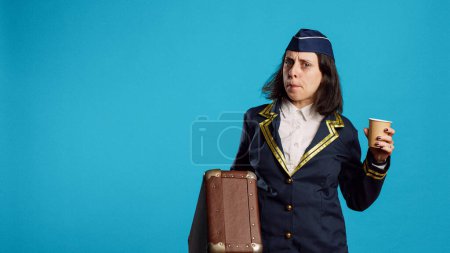 Téléchargez les photos : Smiling air hostess carrying suitcase to leave for work, drinking coffee and getting ready for departure. Stewardess preparing to fly on airplane, holding briefcase and feeling confident. - en image libre de droit