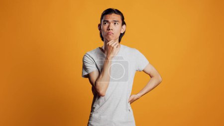 Téléchargez les photos : Pensive young adult thinking about solution in studio, brainstorming ideas to make right decision. Carefree thoughtful model being positive to think about answer, orange background. - en image libre de droit