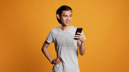 Photo for Modern young adult browsing internet on smartphone, using mobile phone for social media online website in studio. Carefree happy man checking browser app over orange backdrop. - Royalty Free Image