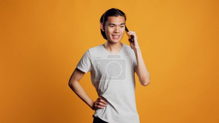Téléchargez les photos : Homme asiatique moderne répondant à un appel téléphonique à la caméra, ayant une conversation à distance sur smartphone. Jeune adulte étant décontracté et insouciant, parlant sur téléphone portable et étant amical. - en image libre de droit