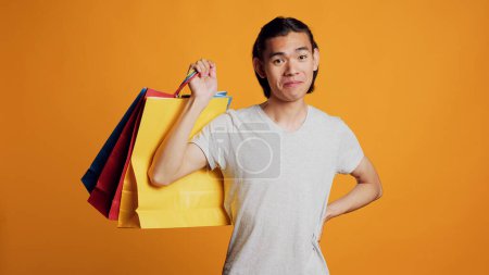 Téléchargez les photos : Asiatique cool homme tenant des sacs à provisions sur fond orange, se sentant heureux et joyeux après avoir acheté des vêtements au centre commercial. Jeune homme portant des sacs en papier et agissant shopaholic sur la caméra. - en image libre de droit