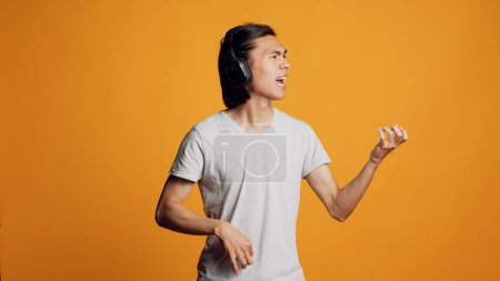 Téléchargez les photos : Happy adult playing air guitar and listening to song, having fun with audio headphones on camera. Male model feeling positive and natural with headset, singing mp3 music and smiling. - en image libre de droit