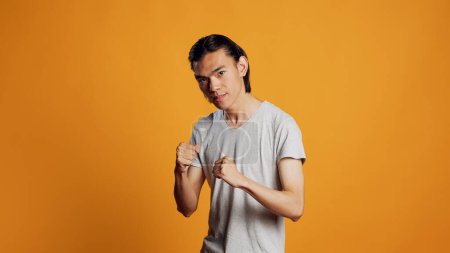 Photo for Asian man clenching fists and acting violent, causing conflict and being aggressive on camera. Young negative person being rebel and punching in studio, angry model on orange backdrop. - Royalty Free Image