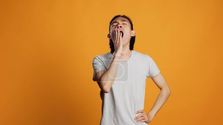 Téléchargez les photos : Drained person yawning and being tired in studio, falling asleep and feeling worn out. Exhausted young man being sleepy and carefree over orange background, having confidence on camera. - en image libre de droit