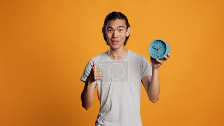 Photo for Smiling model looking at wall clock and feeling good, checking time and hour on watch in studio. Young modern guy feeling happy about being punctual, friendly positive man. Thumbs up. - Royalty Free Image