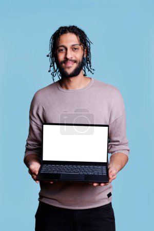 Téléchargez les photos : Un homme arabe souriant tenant un ordinateur portable avec un écran blanc vierge pour la présentation marketing maquette et regardant la caméra. Personne montrant l'appareil portable avec affichage vide pour le logiciel - en image libre de droit