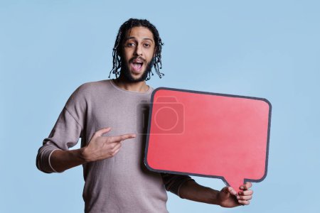 Foto de Hombre árabe excitado sosteniendo la burbuja del habla para anunciar el producto en banner rojo con espacio de copia. Alegre persona sonriente apuntando a la nube de diálogo vacía para el mensaje promocional maqueta - Imagen libre de derechos