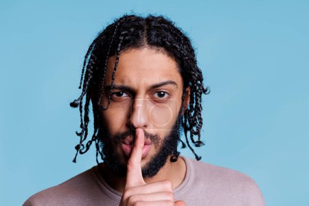 Photo for Arab man making silent gesture with serious facial expression and looking at camera. Young person with raised eyebrow showing quiet sign while holding finger on lips portrait - Royalty Free Image