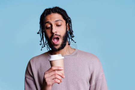 Photo for Surprised arab man holding coffee to go and looking at paper mug lid with shocked facial expression. Confused person drinking take out cappuccino cup while posing for studio shot - Royalty Free Image