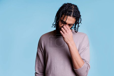 Photo for Tired arab man with closed eyes holding nose while suffering from sinus pain. Exhausted young adult person with black braided hair having headache and fatigue on blue background - Royalty Free Image