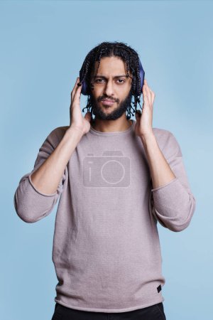 Photo for Cool arab man listening to music while touching headphones on head and looking at camera. Confident person with serious face expression enjoying music in earphone portrait - Royalty Free Image