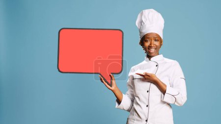 Foto de Mujer joven cocinera sosteniendo burbuja de habla roja en la cámara, posando con cartón copyspace aislado en el estudio. Chef con uniforme de cocina mostrando cartón en blanco en el estudio, servicio de catering. - Imagen libre de derechos