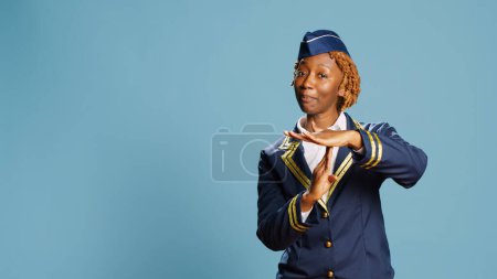 Photo for Young air hostess showing timeout symbol in studio, asking for break and rejecting work. Confident stewardess in flying uniform doing pause and refusal gesture over blue background. - Royalty Free Image