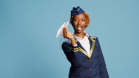 Photo for Female model showing folded airplane in studio, playing with paper plane dressed as stewardess. Professional female airliner in uniform holding origami aircraft on camera over background. - Royalty Free Image