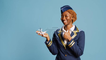 Téléchargez les photos : Hôtesse de l'air souriante tenant un faux petit avion, jouant avec un jouet miniature artificiel en studio. Joyeux avion de ligne heureux s'amuser avec un mini avion d'imitation sur fond bleu. - en image libre de droit