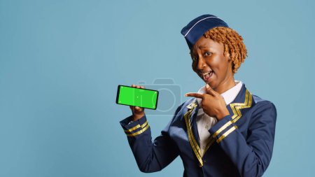 Foto de Chica afroamericana mirando a la pantalla verde en blanco en la cámara, mostrando la pantalla de croma clave aislada en la pantalla del teléfono inteligente. Azafata aérea profesional utilizando copyspace en el teléfono móvil. - Imagen libre de derechos