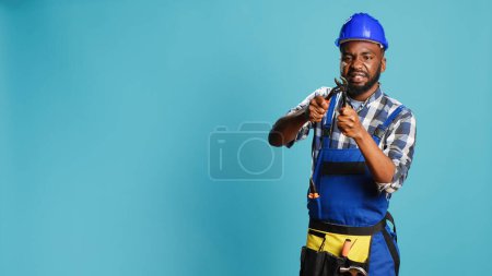 Photo for Professional contractor tightening with wrench, using spanner tool to fix and clamp on camera. Young builder with overalls doing carpentry work in studio, steel instrument key. - Royalty Free Image