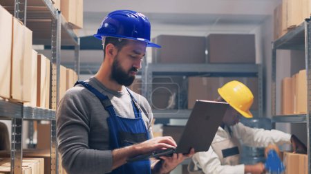 Photo for Young man in overalls reading products list in boxes, planning delivery for industrial service. Employee working with laptop for stock inventory and logistics, business development. Handheld shot. - Royalty Free Image