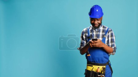 Photo for Construction worker texting messages on smartphone app, using social media browser in studio. Young craftsman browsing online website, wearing building overalls and safety helmet. - Royalty Free Image