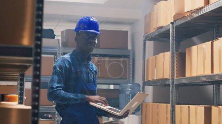 Photo for African american worker counting products on shelves, checking stock list on laptop before shipping order. Male person with hardhat using pc to work on merchandise inventory. Handheld shot. - Royalty Free Image