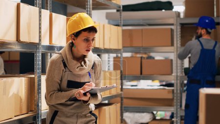 Photo for Female supervisor checking number of merchandise in storage room, counting cardboard boxes with products on depot shelves. Woman using clipboard files to look at logistics list. Handheld shot. - Royalty Free Image