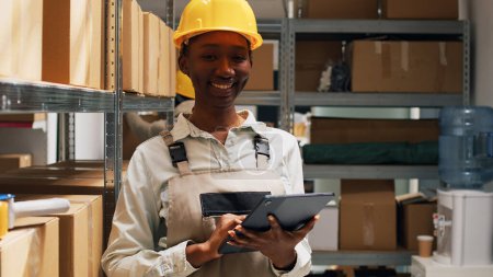 Photo for Female employee in overalls checking tablet inventory, using device to plan logistics to ship merchandise. Young adult checking industrial stock in boxes, package distribution and shipment. - Royalty Free Image