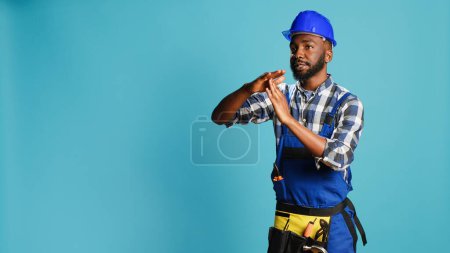 Photo for African american man expressing timeout gesture in studio, asking for break after working on building project. Male builder in overalls showing pause half time symbol on camera. - Royalty Free Image