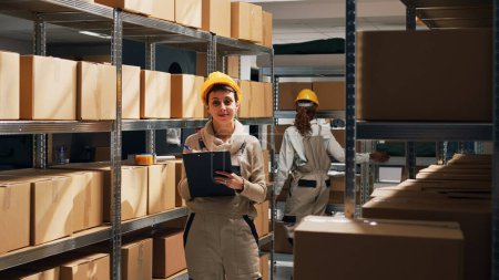 Photo for Depot employee checking stock merchandise on racks in storehouse space, looking at delivery boxes on shelves. Woman working in warehouse and using files for inventory before distribution. - Royalty Free Image