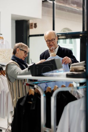 Téléchargez les photos : Couple de personnes âgées regardant chemise à la mode bleue analysant le matériel dans le magasin de vêtements, regardant une nouvelle collection de mode. Personnes âgées qui achètent des vêtements décontractés, l'achat de marchandises à la mode - en image libre de droit